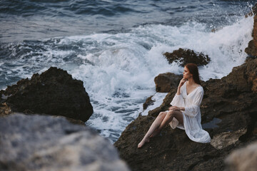 beautiful woman in a white wedding dress sits on the stones by the ocean pensive look