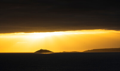 Rays of sun shine through clouds during sunset on a calm winter day with Ballycotton Lighthouse in county Cork, Ireland, in the background