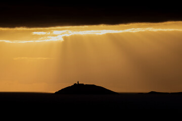 Rays of sun shine through clouds during sunset on a calm winter day with Ballycotton Lighthouse in county Cork, Ireland, in the background