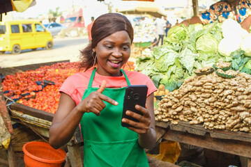 Happy African business woman or female trader wearing a green apron and pointing to a smart phone...