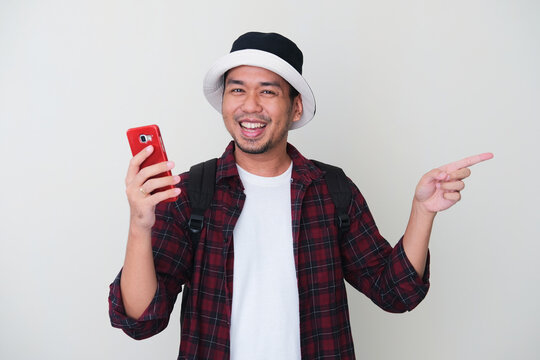 Adult Asian man wearing bucket hat and backpack smiling happy while pointing to his left. Travelling concept