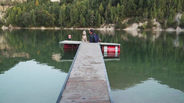 4K Man sitting with his beagle on wooden pier contemplating lake in autumnal season. Nature, environment, animals 4K concept video.