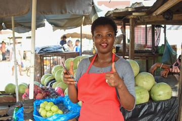 African woman or female trader with a smart phone, standing at her stall in a local market in...