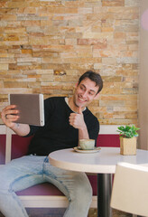 Young and handsome man talking in a videoconference media in a tablet in a coffee shop while having a coffee. High quality photo