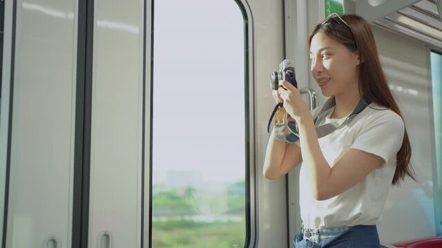 Selective focus of female Asian tourist standing in a running train, taking a picture of the scenery outside the trains through a glass window with a vintage film camera while traveling by sky train.