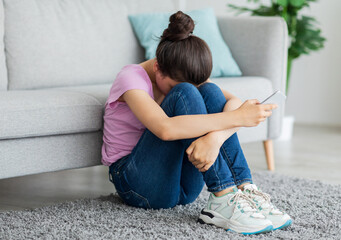 Depressed Indian teen girl with cellphone crying on floor at home, suffering from cyber bullying,...
