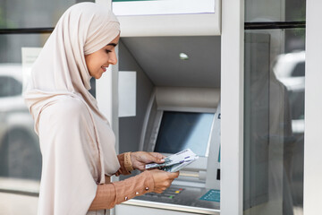 Cheerful millennial arab muslim lady in hijab counts euro near atm with blank screen