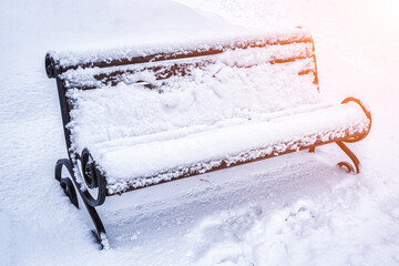 A bench in the park covered with snow and lit by the sun in winter.