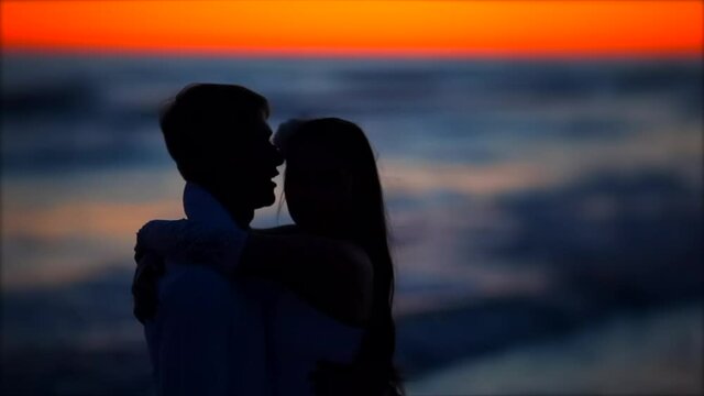 Silhouette of the newlyweds kissing in the evening by the sea against the background of an orange sunset..Young bride and groom walking along the surf line and kissing on wedding day at the sea beach.