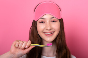 Close-up portrait of caucasian brunette teen girl with freckles in sleep mask holds a toothbrush in hand brushing her teeth morning routine dental hygiene, teeth wide smile isolated on pink background