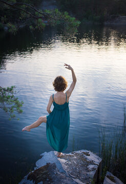 unrecognizable slender girl in a sundress stands with her back on a stone in front of smooth surface of water in a graceful dance pose. Nature. Sunset. Relaxation, enjoying nature, mood to dance