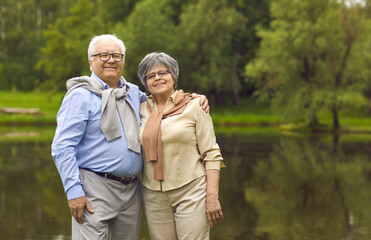 Portrait of smiling elderly pensioners standing in the park by the lake in summer. Senior man and woman are resting outdoors in the park. Happy joyful penionaries having a rest in the park.