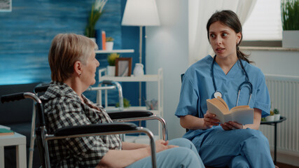 Obraz premium Medical assistant reading book for elder disabled woman in nursing home. Senior patient sitting in wheelchair receiving assistance and support from nurse as entertainment and fun activity.