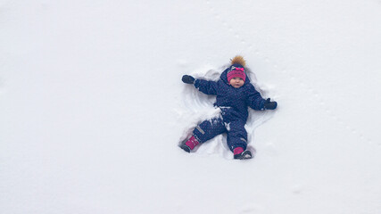 Fototapeta na wymiar A baby girl lies in the snow in winter and waves her hands portraying an angel