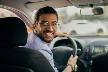 Cheerful middle-eastern taxi driver looking at back seat and smiling - obrazy, fototapety, plakaty