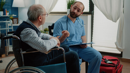 Obraz premium Medical assistant consulting old man with disability in nursing home. Retired patient in wheelchair talking to nurse for checkup about disease and treatment for healthcare. Disabled person