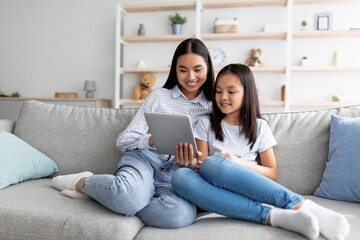 Young asian mother and daughter sitting on sofa and using digital tablet, watching videos or...