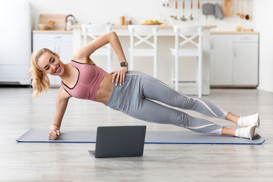 Smiling european young blonde fitness lady doing side plank and watch online workout lesson on laptop