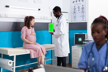 Obraz premium Pregnant patient looking at vertical green screen on tablet in office. Medical physician holding chroma key on device display for mockup template and isolated background at checkup visit.
