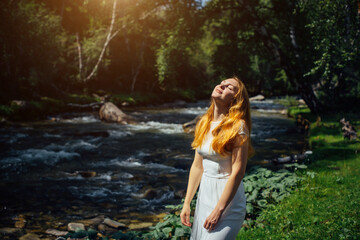 Beautiful woman in white dress with long red hair raised face to the sun on the bank of river in green summer forest.  Portrait of young relaxed girl on the background of nature.