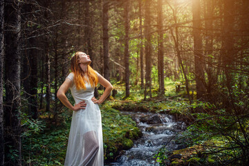 Young woman in white dress raised her face towards the sun. Dreamy pretty girl on the background of green forest and mountain stream.