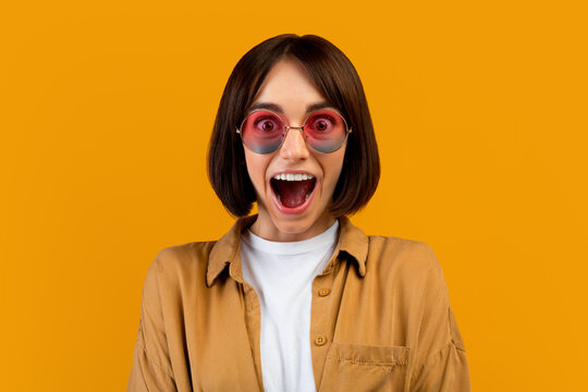 Surprised young woman in colorful sunglasses shouting WOW, opening mouth in excitement over yellow background