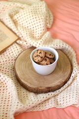Mug filled with chocolate chip cookies, open book, reading glasses and knitted blanket on the bed. Selective focus.