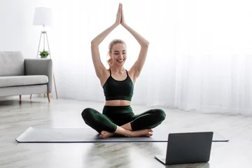 Poster Satisfied caucasian beautiful young blonde woman practicing yoga in lotus position, exercising at home © Prostock-studio