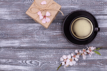 Spring composition with a black cup of coffee, a gift wrapped in kraft paper, spring flowering almond branches with flowers on a gray shabby wooden table. Flat lay, copy space