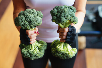 Woman in the gym lifts a broccoli in the form of a dumbbell - obrazy, fototapety, plakaty