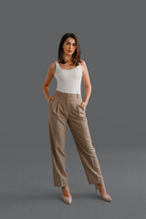 Full length portrait of happy businesswoman looking confident at camera, dressed in white t shirt with short sleeves and beige pants, posing in studio over gray background