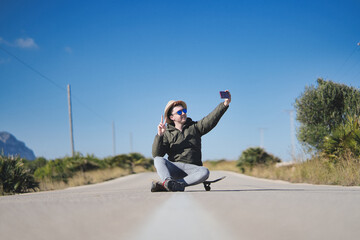 young man with glasses and hat, sitting on a skateboard taking a selfie on a straight street.