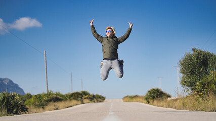 young man with sunglasses and hat, jumping with fingers in symbol of victory. jumping in the middle of a straight road. concept of overcoming and victory.