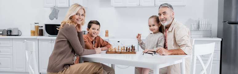 happy kids with grandparents looking at camera while playing chess in kitchen, banner.