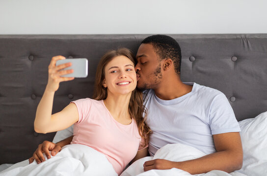 Young diverse couple with cellphone kissing while taking selfie in bed at home