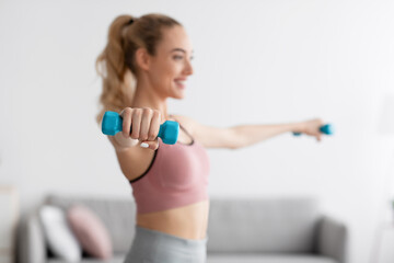 Cheerful european millennial blonde female lifting dumbbells at home, selective focus