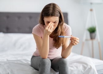 Upset young woman holding pregnancy test, touching her face in stress on bed at home