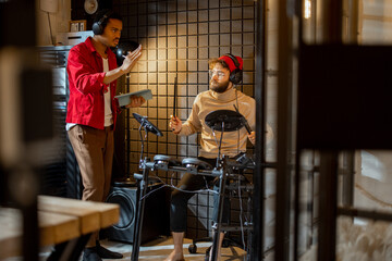 Two stylish men playing electric drums, composing electronic music at small recording studio. Small band creating or practicing music in a home studio. Caucasian and hispanic man playing together