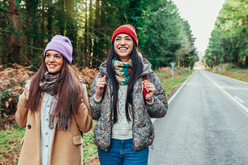 Two cheerful hitchhiker female traveler walking on the roadside with backpack. Two smiling beautiful caucasian sisters hiking on an empty highway. Tourism and travel concept.