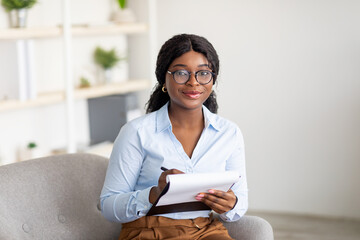 Friendly black female psychologist writing in clipboard, having session with client, sitting on couch at modern office - obrazy, fototapety, plakaty