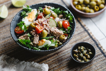 Canned tuna salad with fresh vegetables, capers and olives in a black bowl. Healthy lunch or dinner.