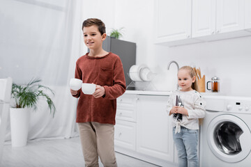 smiling kids holding cups and cutlery in kitchen.