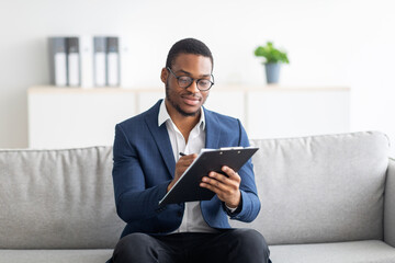 Focused black male psychologist sitting on couch and writing in clipboard at office
