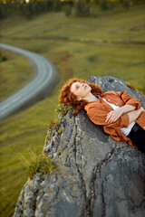 Redhead curly woman is lying on large rock at high altitude. View of nature and greenery in the...