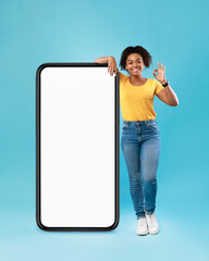 Positive black lady posing near huge cellphone with empty screen, showing okay gesture, recommending website, mockup