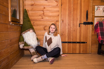 a girl in a winter log cabin sits by the window together with a decorative forest gnome
  