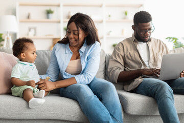 Black mother playing with baby, dad working on pc