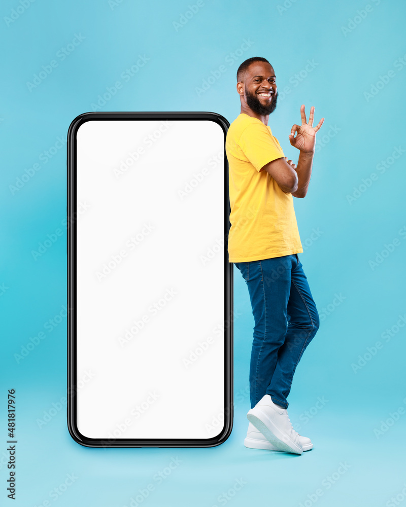 Poster optimistic black man showing ok gesture, leaning on big smarthone with blank screen, recommending ne