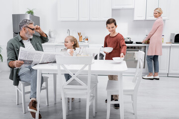 smiling man with newspaper looking at grandchildren setting table for breakfast.