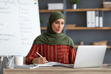 Islamic teacher in headscarf sitting at desk looking at laptop